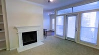 This single-floor space offers a well-designed open layout that seamlessly connects the living and dining areas. The living room is anchored by a classic white-mantled fireplace with a tile surround, adding a focal point of warmth and character to the space. Crown molding runs along the ceiling, lending a refined, polished finish to the room. Soft, neutral carpeting covers the living area floor, contributing to a comfortable and inviting atmosphere.

Natural light is a prominent feature, delivered through a set of full-length French doors paired with transom windows above, allowing generous daylight to fill the space. Blinds on the doors and adjacent window offer privacy and light control. The doors provide direct access to an outdoor area, where greenery is visible through the glass.

Transitioning into the adjacent dining or secondary space, the flooring shifts to a darker hardwood, creating a subtle visual distinction between the two zones. A ceiling fan with a track lighting fixture illuminates this area, while an additional window further enhances brightness. The overall ambiance is clean, neutral, and move-in ready.