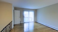 This single-floor residence offers a clean and functional layout designed for comfortable everyday living. The main living area immediately draws attention with its polished hardwood floors that reflect natural light, creating a warm and inviting atmosphere throughout the space. A large sliding glass door fitted with vertical blinds serves as the focal point of the room, offering direct access to an outdoor balcony and allowing generous amounts of natural light to pour in. The neutral-toned walls provide a versatile backdrop, well-suited to a variety of interior styles and furnishing choices. A baseboard heating unit runs along the exterior wall, ensuring efficient climate control during cooler months. A solid white panel door provides access to an adjacent room or closet space, contributing to the overall practicality of the floor plan. The space maintains a straightforward, uncluttered aesthetic throughout, with smooth ceilings and clean lines that give it an open, airy feel. The combination of natural light, hardwood flooring, and outdoor access makes this a bright and functional living space suitable for a range of occupants.