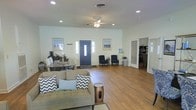 This single-floor space offers an open, airy layout that seamlessly integrates the living and dining areas into one cohesive environment. The interior is finished with warm wood-look flooring that runs throughout, complemented by crisp white walls that enhance the sense of openness and light.

The living area is furnished with a tufted sofa accented by chevron and solid throw pillows, paired with a decorative drum-style side table, creating a relaxed yet styled seating arrangement. Recessed lighting and a ceiling fan with an integrated light fixture provide both ambient illumination and practical airflow. Coastal-themed framed artwork adorns the walls, reinforcing a calm, neutral aesthetic.

Adjacent to the living space, a dining area features a wooden table with seating for multiple guests, positioned near a coffee station, making the space well-suited for both casual meals and gatherings. A bold navy blue front entry door serves as a focal point, flanked by sidelight windows that allow natural light to filter in.

Toward the rear, a seating nook with two dark armchairs offers a quieter corner, while a louvered door leads to an additional room visible through plantation shutters. The overall ambiance is clean, functional, and inviting, with a consistent coastal-casual design theme carried throughout.