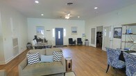 This single-floor space offers an open, airy layout that seamlessly integrates the living and dining areas into one cohesive environment. The interior is finished with warm wood-look flooring that runs throughout, complemented by crisp white walls that enhance the sense of openness and light.

The living area is furnished with a tufted sofa accented by chevron and solid throw pillows, paired with a decorative drum-style side table, creating a relaxed yet styled seating arrangement. Recessed lighting and a ceiling fan with an integrated light fixture provide both ambient illumination and practical airflow. Coastal-themed framed artwork adorns the walls, reinforcing a calm, neutral aesthetic.

Adjacent to the living space, a dining area features a wooden table with seating for multiple guests, positioned near a coffee station, making the space well-suited for both casual meals and gatherings. A bold navy blue front entry door serves as a focal point, flanked by sidelight windows that allow natural light to filter in.

Toward the rear, a seating nook with two dark armchairs offers a quieter corner, while a louvered door leads to an additional room visible through plantation shutters. The overall ambiance is clean, functional, and inviting, with a consistent coastal-casual design theme carried throughout.