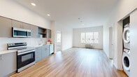 This single-floor unit showcases a clean, contemporary design with a practical and open layout. The kitchen is a standout feature, equipped with a full suite of stainless steel appliances including an electric range, over-the-range microwave, dishwasher, and refrigerator. Sleek gray cabinetry lines the walls, complemented by white countertops and recessed lighting overhead, creating a bright and functional cooking space. The kitchen flows seamlessly into the open living area, which is finished with warm-toned hardwood-style flooring that extends throughout the main spaces, adding continuity and a sense of openness.

A dedicated living zone benefits from a large window that draws in natural light and offers exterior views, with a wall-mounted PTAC unit providing climate control. The bedroom areas are carpeted for comfort and similarly receive natural light through sliding windows with outdoor views. The neutral white walls and clean lines throughout the unit contribute to a bright, minimalist aesthetic. Additional cabinetry is tucked thoughtfully into transitional spaces, offering practical storage without disrupting the streamlined character of the interior.