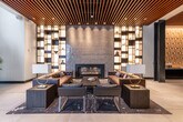 This single-floor space functions as a refined residential or hospitality lobby, unified by a striking slatted wood ceiling that runs throughout and recessed lighting that creates a warm, layered ambiance across all zones. Large-format porcelain tile flooring in a neutral tone anchors the open layout, reflecting light and reinforcing the sense of generous, open space.

The main lounge area is anchored by a sleek linear gas fireplace set against a textured stone accent wall, flanked on both sides by floor-to-ceiling illuminated shelving units displaying curated decorative objects. Deep leather sofas and low-profile chairs are arranged around a dark stone coffee table atop a patterned area rug, creating an inviting and sophisticated seating environment. Floor-to-ceiling glazing at the entrance allows natural light to pour in and provides visibility to the street beyond.

The reception area features a substantial live-edge wood desk with matte black pendant lighting overhead, attended by staff. Framed black-and-white photography and potted fiddle-leaf fig trees add warmth to the otherwise clean, contemporary aesthetic.

A separate lounge and bar area includes communal high-top tables, bar seating, a wood-and-concrete service counter, and additional illuminated shelving, making it well-suited for social gatherings or casual work settings.