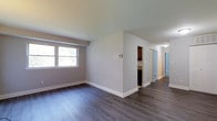 This single-floor space presents a clean, modern interior defined by dark hardwood-style flooring that runs consistently throughout, creating a cohesive and polished look. The main living area is finished with smooth, neutral grey walls and crisp white baseboards, contributing to a bright and contemporary aesthetic. A window along one wall allows natural light to filter into the space, enhancing the open feel of the room.

A partial wall divides the main living area from the rest of the unit, where a corridor leads toward additional rooms. Visible from the main area, the kitchen features dark cabinetry and appears to connect naturally to the hallway. The hallway extends toward what appears to be at least one additional room or bathroom, suggested by a light-colored door at the far end.

The layout is straightforward and functional, with an efficient use of space throughout. Ceiling-mounted lighting is visible in the hallway, complementing the natural light from the main room's window. The overall ambiance is clean and move-in ready, with modern finishes that provide a neutral canvas suitable for a variety of interior styles.