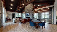 This impressive single-floor gathering space blends rustic elegance with modern comfort through a thoughtfully curated design. The centerpiece of the main lounge is a dramatic floor-to-ceiling stone fireplace wall, topped with a wall-mounted television and flanked by rich leather armchairs and a plush sectional sofa arranged over a patterned area rug. Soaring vaulted ceilings with exposed dark-stained timber trusses and beams add striking architectural character, while recessed lighting and stylish sconces provide warm, layered illumination throughout.

Large-paned windows and glass wall partitions allow natural light to flood the interior, offering views of the surrounding greenery and an adjacent outdoor area. Light hardwood flooring runs continuously throughout, unifying the open-plan layout. A dedicated games zone features a round poker table surrounded by navy upholstered chairs, and a separate wing houses a full-size billiards table.

The bar and kitchen area is anchored by an oversized granite island with tufted counter stools, pendant lighting, dark cabinetry, and an exposed dark brick accent wall — ideal for casual dining or entertaining. A dining high-top table with additional seating sits near the glass-panel walls, completing this well-appointed, versatile space.