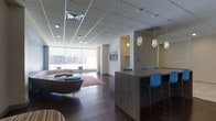 This single-floor space showcases a thoughtfully designed open-plan layout that seamlessly integrates a lounge area and a bar-height dining counter. The lounge is anchored by a curved sectional sofa in warm taupe tones, complemented by a low ottoman and a vibrant area rug that adds a pop of color to the rich dark hardwood flooring running throughout the space. A wall-mounted flat-screen television serves as a focal point in the central partition, creating a subtle division between the seating and dining zones without interrupting the open feel.

The bar-height counter, finished in a natural wood-tone laminate, is lined with sleek blue chairs that introduce a bold accent color against the neutral palette. Above, a cluster of decorative pendant lights with ornate glass shades adds warmth and visual interest to the dining area. The drop ceiling with recessed lighting ensures even illumination throughout.

Large floor-to-ceiling windows along one wall flood the lounge with abundant natural light and offer open views of the surrounding urban landscape. The overall ambiance is modern and inviting, balancing functionality with contemporary design sensibility.