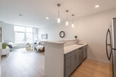 This thoughtfully designed single-floor residence combines modern aesthetics with functional living. The open-concept layout seamlessly connects the kitchen and living area, creating a bright and airy atmosphere. The kitchen features sleek gray cabinetry, quartz countertops, stainless steel appliances including a refrigerator, range, and built-in microwave, and a peninsula with pendant lighting that doubles as a casual dining or entertaining space. Light wood-tone flooring runs throughout the main living areas, enhancing the clean, contemporary feel.

The living room is a standout highlight, anchored by floor-to-ceiling windows that flood the space with natural light and offer views of the surrounding streetscape. A vibrant abstract painting adds a pop of color against the neutral walls, complementing the modern furnishings.

The bedroom is a serene retreat, featuring plush carpeting, dual windows with street views, and a bold teal accent wall that adds depth and character. A walk-in closet provides generous storage. The bathroom is clean and well-appointed, with a gray vanity, quartz countertop, and light wood-tone flooring that ties back to the rest of the home. Recessed lighting and a well-placed vanity mirror complete the polished look throughout this stylish residence.