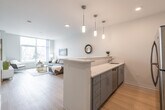 This thoughtfully designed single-floor residence combines modern aesthetics with functional living. The open-concept layout seamlessly connects the kitchen and living area, creating a bright and airy atmosphere. The kitchen features sleek gray cabinetry, quartz countertops, stainless steel appliances including a refrigerator, range, and built-in microwave, and a peninsula with pendant lighting that doubles as a casual dining or entertaining space. Light wood-tone flooring runs throughout the main living areas, enhancing the clean, contemporary feel.

The living room is a standout highlight, anchored by floor-to-ceiling windows that flood the space with natural light and offer views of the surrounding streetscape. A vibrant abstract painting adds a pop of color against the neutral walls, complementing the modern furnishings.

The bedroom is a serene retreat, featuring plush carpeting, dual windows with street views, and a bold teal accent wall that adds depth and character. A walk-in closet provides generous storage. The bathroom is clean and well-appointed, with a gray vanity, quartz countertop, and light wood-tone flooring that ties back to the rest of the home. Recessed lighting and a well-placed vanity mirror complete the polished look throughout this stylish residence.