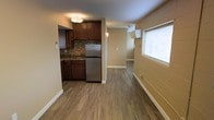 This single-floor unit offers an efficient and practical layout with a seamless flow between its main living areas. The kitchen is a standout feature, equipped with rich dark wood cabinetry, a mosaic tile backsplash, dark stone countertops, and a stainless steel refrigerator. A flush-mount ceiling light illuminates the kitchen area, and a sink is positioned beneath the upper cabinets, maximizing counter space. The flooring throughout the unit consists of durable hardwood-style planks in a warm medium-brown tone, providing a cohesive look across all rooms. A hallway extends from the kitchen area toward additional rooms, lined with baseboard heating units that run along the walls. Natural light enters through a window fitted with white blinds, brightening the living space. The walls are finished in a neutral beige tone, complementing the overall warm color palette. A wall-mounted unit, likely for heating or cooling, is visible further down the hallway, adding to the unit's functional amenities. The space presents a straightforward, no-frills design focused on practicality and utility.