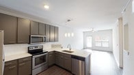 This single-floor unit is designed with a clean, contemporary aesthetic that emphasizes open living and functional design. The kitchen serves as a central highlight, featuring gray flat-front cabinetry extending to the ceiling, a white subway tile backsplash, light quartz countertops, and a full suite of stainless steel appliances including a range, microwave, dishwasher, and refrigerator. A curved peninsula with an integrated sink and pendant lighting above creates a natural transition into the open living area, making the space feel connected and sociable.

The living area is generously sized, finished with warm hardwood-style flooring, crisp white walls, and a ceiling fan. Large windows allow natural light to pour in, and a glass-panel door provides additional brightness and outdoor access.

The bedroom continues the neutral tone with plush carpeting, a ceiling fan, and dual windows that bring in ample natural light while offering views of the surrounding area. The bathroom is finished with white subway tile surrounding a soaking tub and shower combination, complemented by recessed lighting and wood-style flooring. Overall, the unit presents a cohesive, modern feel with durable, well-chosen finishes throughout.