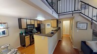 This two-story residence offers a well-organized layout across both levels, with distinct living, sleeping, and functional spaces. The main level features a galley-style kitchen equipped with light wood cabinetry, dark granite-style countertops, stainless steel appliances including a refrigerator and microwave, a coil-top range, and a double stainless sink. Wood-look flooring runs through the kitchen and extends into the adjacent hallway, adding warmth to the lower level.

The living room is a standout area, anchored by a tile-surround fireplace and complemented by vaulted ceilings and a sliding glass door that brings in natural light and outdoor views. A carpeted staircase with a black metal railing leads to the upper level, where an open loft area provides flexible living space with tall angled walls and carpeted flooring, suitable for a sitting room or home office.

The bedroom features neutral-toned walls, carpeted floors, and a window overlooking greenery outdoors. The bathroom includes a full soaking tub with a shower combination and chrome fixtures, finished in clean, neutral tones throughout.