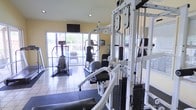 This single-floor fitness center is designed to support a comprehensive workout experience. The space is outfitted with a robust multi-station cable weight machine for strength training, alongside a selection of cardio equipment including stair climbers and an elliptical trainer. Light-colored tile flooring covers much of the room, complemented by darker rubber matting in the training areas to provide stability and cushioning during exercise. The walls are painted in a warm yellow tone, creating an energetic yet welcoming atmosphere. Recessed lighting in the ceiling, combined with abundant natural light streaming through large sliding glass doors and adjacent windows, keeps the space bright and airy. The glass doors open to an outdoor patio or balcony area, offering views of greenery beyond and a refreshing connection to the outdoors. A wall-mounted flat-screen television is positioned for visibility across the room, adding an entertainment option during workouts. The overall layout is open and functional, allowing for comfortable movement between equipment while maintaining an organized and motivating environment suitable for a range of fitness routines.