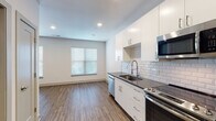 Located at 237 W 4th St in Kansas City, MO, this single-floor unit offers a clean, modern aesthetic throughout. The kitchen is a standout feature, equipped with sleek white cabinetry, dark countertops, a white subway tile backsplash, and a full suite of stainless steel appliances including a refrigerator, electric range, built-in microwave, and dishwasher. A professional-style spring-neck faucet adds a refined touch to the undermount sink. The open layout connects the kitchen seamlessly to the living area, which is bright and airy, benefiting from natural light through a window with blinds. Durable wood-look plank flooring runs consistently throughout the unit, lending warmth and cohesion to each space. The bedroom is generously sized with two windows that allow ample natural light to fill the room, complemented by neutral gray walls and the same hardwood-style flooring. The bathroom is finished to a high standard, featuring dark countertops with an undermount sink, a backlit vanity mirror, white subway tile surrounding the tub/shower combination, and brushed nickel fixtures and grab bars. Recessed lighting throughout the unit provides a clean, polished look. This thoughtfully designed unit offers a contemporary living experience in Kansas City.