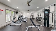 This fitness center offers a thoughtfully designed workout space spread across a single open floor. The interior features crisp white walls, recessed lighting, and durable speckled rubber flooring throughout, creating a clean and functional environment well-suited for a variety of exercise routines.

The cardio zone is equipped with multiple treadmills, elliptical trainers, and a stationary bike, all positioned along one side of the room to maximize open floor space. A wall-mounted television is conveniently placed for viewing during workouts. On the opposite side, a full dumbbell rack stocked with a wide range of weights is paired with a multi-use cable machine and adjustable weight benches, catering to both strength and resistance training needs.

Two ceiling fans are installed overhead, complementing the wall-mounted air conditioning unit to ensure proper climate control and airflow during intense workouts. Natural light filters in through multiple windows along the walls, contributing to a bright and energizing atmosphere. Large mirrors line one wall, enhancing the sense of space and allowing users to monitor their form. A dedicated entry door with windows provides additional natural light and easy access. The overall layout is efficient and well-organized, making the space accessible and practical for a broad range of fitness activities.