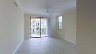 This single-floor space showcases a light-filled interior defined by neutral, warm-toned walls and large-format tile flooring that extends throughout the main living area. The room is bathed in natural light, courtesy of a sliding glass door that opens to a private balcony with a wrought iron railing, offering views of lush tropical landscaping and the surrounding Mediterranean-style architecture. A secondary window fitted with horizontal blinds provides additional natural light and ventilation. Crown molding runs along the ceiling perimeter, lending a refined architectural detail to the space, while clean white baseboards frame the flooring below. A ceiling fan positioned at the center of the room offers practical climate control and adds to the airy, open atmosphere. The overall ambiance is calm and bright, with the combination of soft wall tones, natural light, and outdoor greenery creating a welcoming, comfortable environment well-suited to a warm-weather setting.