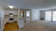 This single-floor unit offers an open-concept design that seamlessly connects the kitchen and living area, creating a spacious and functional layout. The kitchen is outfitted with white cabinetry, a built-in microwave, an electric range, and a peninsula that serves as both a prep area and a divider between the kitchen and the carpeted living space. The kitchen features hardwood flooring, while the living and bedroom areas are finished with neutral carpet, providing a comfortable underfoot feel throughout. A wall-mounted air conditioning unit is visible in the living area, complementing the baseboard heating system found throughout the unit. A doorway from the living area leads into a bedroom, which benefits from natural light courtesy of a window. The bedroom also features its own wall unit for climate control, ensuring independent temperature management. The overall color palette is light and neutral, with white walls and trim throughout, contributing to a clean, bright atmosphere. The layout is straightforward and efficient, maximizing usable space while maintaining an open, airy feel across the main living areas.