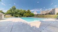 This outdoor community pool facility is thoughtfully designed across two levels, offering a well-appointed recreational space for residents and guests. The centerpiece is a large, rectangular swimming pool with crystal-clear turquoise water, accessible via stainless steel entry ladders. A lifeguard stand positioned poolside underscores the facility's commitment to safety and supervision.

The surrounding deck is generously proportioned, providing ample space for movement and relaxation. Along the perimeter, a long row of reclining lounge chairs is neatly arranged, accompanied by teal patio umbrellas that offer shade on sunny days. The entire pool area is enclosed by a sturdy wrought iron fence, ensuring security and clear boundaries.

A dedicated pool house structure sits adjacent to the water, offering a practical support facility for the space. A second building with a steep gabled roofline and exterior staircase is also present, adding architectural character to the setting. Mature trees and well-maintained greenery surround the enclosure, creating a naturally sheltered atmosphere. Views of neighboring multi-story residential buildings are visible in the background, situating this amenity within a larger community context. The space benefits from abundant natural light and an open-sky setting, making it an inviting outdoor retreat.