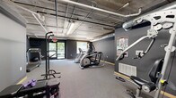 This fitness center is designed with a utilitarian yet purposeful aesthetic, making it a practical space for a comprehensive workout. The room is finished with dark charcoal-gray walls and neutral carpet flooring, creating a focused, gym-like atmosphere throughout. An exposed concrete ceiling with visible pipework, ductwork, and fluorescent strip lighting gives the space a distinct industrial character.

The fitness center is divided into clearly defined workout zones. The cardio area is equipped with a treadmill, an elliptical cross-trainer, and a stair-stepper machine, all positioned to take advantage of natural light from large windows and a glass door that provide views of the surrounding greenery. The strength training area features a multi-function cable machine, a power tower for pull-ups and dips, a weight bench, and a set of dumbbells, catering to a variety of resistance training exercises. A fitness reference poster is mounted on the wall for exercise guidance.

The layout of the space allows for free movement between equipment, and the carpeted floor provides comfort underfoot. Ventilation is handled by exposed HVAC ductwork integrated into the ceiling. The space is accessed through a dedicated entry door and benefits from both artificial and natural light sources, ensuring a bright and well-ventilated environment for exercise.