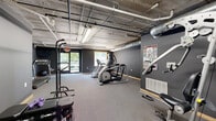 This fitness center is designed with a utilitarian yet purposeful aesthetic, making it a practical space for a comprehensive workout. The room is finished with dark charcoal-gray walls and neutral carpet flooring, creating a focused, gym-like atmosphere throughout. An exposed concrete ceiling with visible pipework, ductwork, and fluorescent strip lighting gives the space a distinct industrial character.

The fitness center is divided into clearly defined workout zones. The cardio area is equipped with a treadmill, an elliptical cross-trainer, and a stair-stepper machine, all positioned to take advantage of natural light from large windows and a glass door that provide views of the surrounding greenery. The strength training area features a multi-function cable machine, a power tower for pull-ups and dips, a weight bench, and a set of dumbbells, catering to a variety of resistance training exercises. A fitness reference poster is mounted on the wall for exercise guidance.

The layout of the space allows for free movement between equipment, and the carpeted floor provides comfort underfoot. Ventilation is handled by exposed HVAC ductwork integrated into the ceiling. The space is accessed through a dedicated entry door and benefits from both artificial and natural light sources, ensuring a bright and well-ventilated environment for exercise.