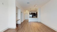 This single-floor space showcases a thoughtfully designed open-concept layout that seamlessly connects the living area and kitchen, creating a bright and airy atmosphere. The warm hardwood-style flooring flows continuously throughout the main living space, adding a sense of warmth and cohesion to the interior.

The kitchen is a standout feature, equipped with dark cabinetry that provides a striking contrast against the white countertops and light walls. A peninsula with a built-in sink offers both additional prep space and a natural division between the kitchen and living area. Stainless steel appliances, including a refrigerator and range, add a sleek, modern touch. Pendant lighting hangs above the peninsula, complementing the track lighting overhead that illuminates the kitchen with focused, adjustable light.

The living area is generously proportioned, offering a versatile open canvas with crisp white walls and clean architectural lines. A built-in ledge along one wall provides a functional design element. The entryway features a full-panel door and is integrated smoothly into the overall floor plan. Throughout the space, the combination of contemporary finishes, neutral tones, and efficient layout makes for a functional and visually appealing interior.