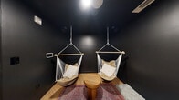 This single-floor wellness suite is thoughtfully designed to support contrast therapy and full-body recovery, comprising four distinct zones within a compact footprint. The main reception-style area features striking white subway-tiled walls extending from floor to mid-wall, paired with a deep matte black ceiling that creates a bold, high-contrast aesthetic. Framed informational signage on the "Hot, Cold, Rest, Repeat" regimen lines the walls, accompanied by a white open-cube storage unit with woven basket drawers and a potted ZZ plant. A glass-paneled black door connects this space to the broader facility. The dedicated massage and meditation room is finished in an enveloping matte black, with warm wood-tone flooring and two hanging macramé hammock chairs suspended from the ceiling, creating an intimate and serene retreat. A patterned area rug, a solid wood side table, and floor cushions complete the space. The traditional cedar sauna features full tongue-and-groove wood paneling on all walls, ceiling, and tiered benches, evoking an authentic Finnish sauna experience. An accessible shower stall rounds out the suite, finished in clean white with stainless steel grab bars, a hand-held shower head, and a teak fold-down bench for comfort and safety.