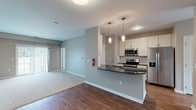 This well-appointed single-floor residence offers a thoughtfully designed open-concept layout that seamlessly connects the main living and dining areas to the kitchen. The kitchen is a standout feature, showcasing a peninsula with dark granite countertops, white shaker-style cabinetry, a classic subway tile backsplash, stainless steel appliances including a range and microwave, and stylish pendant lighting overhead. Rich hardwood-style flooring runs through the kitchen and main living corridor, while plush carpeting adds comfort to the bedrooms and living area. Neutral gray tones on the walls create a clean, contemporary backdrop throughout. The bedrooms are generously sized, each featuring large windows that invite abundant natural light and views of the surrounding greenery. The primary bathroom includes a dual-sink vanity with dark countertops, hardwood-style flooring, and access to a walk-in closet. A second bathroom offers a full soaking tub and shower combination. A dedicated utility room houses the HVAC system, water heater, and in-unit washer and dryer connections, providing practical convenience. The overall design balances modern finishes with functional living spaces, making this a comfortable and cohesive home.