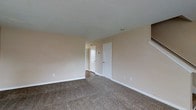 This two-story space is designed with a straightforward, functional layout that maximizes usability. The main living area features wall-to-wall carpeting in a neutral tone, providing a comfortable and cohesive feel underfoot. The walls are finished in a soft beige, creating a warm and understated backdrop throughout. A textured popcorn ceiling adds a subtle visual detail overhead.

The room benefits from a clean, open layout with white-trimmed doors and baseboards providing crisp contrast against the neutral walls. Multiple doorways are visible, suggesting access to additional rooms or hallways, enhancing the overall flow of the space. A corridor leads further into the unit, hinting at additional rooms beyond the main living area. The staircase, partially visible, connects to the second floor, offering additional living space above.

Electrical outlets are conveniently positioned along the walls, and a visible vent ensures climate control throughout. The overall ambiance is practical and move-in ready, with neutral finishes that offer flexibility for a variety of furnishing and décor styles.