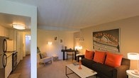 This single-floor living space offers a comfortable and well-appointed layout centered around a carpeted living area. The room is finished with neutral-toned walls and a textured ceiling, creating a warm and understated backdrop throughout. The living area is furnished with a dark sofa accented by vibrant orange throw pillows, paired with a sleek rectangular coffee table that anchors the seating arrangement. A floor lamp and table lamp provide soft, ambient lighting, contributing to the room's cozy atmosphere. A wicker accent chair with a patterned cushion adds a casual, relaxed touch to one corner of the space. Along the back wall, a compact desk and chair serve as a dedicated workspace, flanked by framed artwork and small decorative shelving. A bold graphic print of a Union Station sign serves as a striking focal point on the far wall, adding a urban, stylistic character to the room. The space flows naturally and makes efficient use of its footprint, balancing functional zones for relaxing and working within a single, open area. Overall, the interior presents a tidy, modern-casual aesthetic with warm lighting and thoughtful decorative touches.