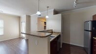 This single-floor residence offers a well-designed layout with consistent finishes throughout. The kitchen serves as a central highlight, featuring a wraparound granite countertop peninsula with pendant lighting overhead, dark wood cabinetry, and stainless steel appliances including a refrigerator, range, and built-in microwave. A secondary sink is integrated into the main counter run, providing added functionality. Crown molding accents the kitchen and living areas, lending an elevated, polished look to the space.

Rich hardwood-style flooring flows continuously through the main living areas, bedrooms, and bathroom, creating a cohesive and warm aesthetic. The living area is generously proportioned, featuring crown molding, a ceiling fan with light fixture, and multiple windows fitted with horizontal blinds that allow for ample natural light. A sliding glass door provides additional light and access to an outdoor area.

The bedroom offers a comfortable footprint with a large window and ceiling fan, while a second room provides flexible use with its own window and pendant light fixture. The bathroom is cleanly appointed with a full tub and tile surround, a dark wood vanity with a light countertop, and matching hardwood-style flooring. An entry area with a security keypad panel completes this functional and neatly finished residence.