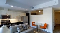 This single-floor unit features an open-concept layout that seamlessly combines the living, dining, and kitchen areas into one cohesive space. The kitchen is outfitted with white shaker-style cabinetry, subway tile backsplash, a stainless steel refrigerator, and a undermount sink — offering a clean, modern aesthetic. Recessed lighting and a ceiling-mounted HVAC vent contribute to a well-lit and climate-controlled environment.

Adjacent to the kitchen, a compact dining area accommodates a round glass-top table paired with two vibrant orange chairs, adding a bold pop of color against the neutral white walls and light wood-tone flooring. A small pendant light fixture overhead defines the dining zone within the open space.

The living area features a gray upholstered sofa dressed with patterned throw pillows, creating a comfortable and inviting atmosphere. A partial wall with a rectangular cutout subtly separates visual zones without fully enclosing the space, maintaining an airy, open feel throughout. The combination of neutral tones, modern fixtures, and warm accent colors gives the unit a practical yet stylish character.