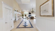 Located at 3895 Waterside Dr in Elizabeth City, NC, this elegantly appointed single-story clubhouse offers a generously sized, open-concept layout filled with natural light and coastal charm. The grand entrance hallway sets the tone with soaring two-story ceilings, large-format cream tile flooring accented by a diamond inlay pattern, white beadboard wainscoting, decorative wall art, and a statement chandelier. Floor-to-ceiling glass doors at the end of the hall frame a bright, welcoming entry.

The expansive great room flows seamlessly from the hallway and features dramatic vaulted ceilings, a striking white-trimmed fireplace flanked by large windows and French doors that open to an outdoor pool and waterfront area. A wall-mounted television above the fireplace and coastal nautical décor — including model sailboats displayed on a high shelf — reinforce the waterside theme throughout.

Adjacent to the main seating area, a pergola-style ceiling defines a casual kitchenette and bar space with warm wood cabinetry, dark countertops, a mounted TV, and wicker bar seating — all surrounded by windows overlooking lush greenery. The entire space is tied together with neutral warm tones, natural fiber rugs, and elegant chandelier lighting, creating a refined yet relaxed coastal atmosphere.