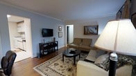 This single-floor space centers around a well-appointed living room that combines classic architectural detailing with a warm, inviting atmosphere. Crown molding lines the ceiling, adding a refined touch to the soft blue-gray walls that lend the room a calm, neutral tone. Hardwood floors run throughout, contributing warmth and a sense of continuity to the space.

The living room is thoughtfully arranged to maximize comfort and function. Seating includes a sofa and a pair of tufted armchairs in complementary neutral tones, positioned around a round dark-wood coffee table atop a patterned area rug with floral detailing. A wall-mounted flat-screen television is supported by a dark wood media console along one wall, which provides additional storage and display space.

Decorative framed artwork, including a nautical sailing scene, adds personality and visual interest to the walls. A floor lamp provides warm ambient lighting to the seating area. A doorway visible from the living room suggests additional rooms beyond, indicating further living or sleeping areas within the space. Overall, the space presents a classic, tidy, and livable environment.