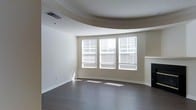 This single-floor space showcases a thoughtfully designed living area highlighted by several architectural details. The ceiling is a standout feature, incorporating a curved tray design that adds depth and visual interest to the room. Crown molding traces the perimeter, complementing the tray ceiling and lending a refined, finished look to the space.

The flooring consists of dark hardwood, which contrasts cleanly with the neutral, warm beige tones of the walls and creates a grounded, cohesive aesthetic throughout. A trio of windows arranged in a bay-style configuration dominates one wall, fitted with white horizontal blinds that allow for adjustable natural light. These windows allow sunlight to pour in, casting bright patterns across the floor and contributing to an open, airy atmosphere.

A fireplace with a clean white mantel is positioned to one side of the windows, serving as a subtle focal point that adds warmth and character to the room. The overall layout feels spacious and open, with clean lines and minimal ornamentation that give the space a versatile, move-in-ready quality suited to a variety of interior styles.