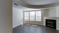 This single-floor space showcases a thoughtfully designed living area highlighted by several architectural details. The ceiling is a standout feature, incorporating a curved tray design that adds depth and visual interest to the room. Crown molding traces the perimeter, complementing the tray ceiling and lending a refined, finished look to the space.

The flooring consists of dark hardwood, which contrasts cleanly with the neutral, warm beige tones of the walls and creates a grounded, cohesive aesthetic throughout. A trio of windows arranged in a bay-style configuration dominates one wall, fitted with white horizontal blinds that allow for adjustable natural light. These windows allow sunlight to pour in, casting bright patterns across the floor and contributing to an open, airy atmosphere.

A fireplace with a clean white mantel is positioned to one side of the windows, serving as a subtle focal point that adds warmth and character to the room. The overall layout feels spacious and open, with clean lines and minimal ornamentation that give the space a versatile, move-in-ready quality suited to a variety of interior styles.