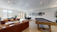 This well-appointed single-floor amenity lounge is designed to serve as a versatile gathering and recreation space. The layout seamlessly integrates multiple functional zones within an open, airy floor plan finished with polished concrete floors and crisp white walls that maximize the sense of brightness throughout.

At the heart of the lounge, a striking floor-to-ceiling natural stone accent wall anchors a linear gas fireplace, with a large flat-screen television mounted above — creating a warm and inviting focal point. Rich cognac leather sofas arranged around a reclaimed wood coffee table and a cowhide area rug provide generous, comfortable seating in this central living area.

Adjacent to the lounge seating, a dedicated game area features a solid wood billiards table, offering a standout recreational amenity. A cozy dining nook nearby includes a round marble-topped table surrounded by modern upholstered chairs, set atop a natural jute rug. A curated gallery wall of black-and-white framed photographs adds a sophisticated, artistic touch to the space.

Natural light flows in through large sliding glass doors and French doors with warm wood framing, offering views of and access to an outdoor area. Tall indoor plants are strategically placed throughout, adding organic warmth to the contemporary interior. Recessed lighting provides even illumination, rounding out this thoughtfully designed communal space.
