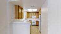 This single-floor residence offers a thoughtfully laid-out living environment anchored by a well-appointed kitchen. The kitchen is a standout feature, designed in a U-shaped configuration that maximizes storage and workflow efficiency. Warm honey-toned wood cabinetry lines the walls from counter to ceiling, providing generous storage space, while light-colored countertops offer ample workspace. A peninsula extends outward, creating a natural divide between the kitchen and adjacent living areas, and serving as an ideal spot for casual dining or food preparation. Natural light fills the kitchen through a window above the sink and a large skylight panel recessed into the ceiling, brightening the space throughout the day. White appliances — including a gas range, over-the-range microwave, and full-size refrigerator — complement the clean, neutral aesthetic of the room. Light-toned tile flooring ties the space together, adding a sense of continuity and ease of maintenance. The overall ambiance of this residence is clean, practical, and welcoming, with a design that prioritizes functionality while maintaining a comfortable, lived-in warmth suitable for everyday living.