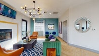 This single-floor community amenity space is thoughtfully designed to encourage socializing and recreation. The lounge area makes an immediate impression with its bold teal accent wall, a striking geometric chandelier with Edison-style bulbs, and a cozy fireplace framed in a yellow-bordered patterned tile surround. Seating is both stylish and inviting, featuring a plush green velvet sofa, cognac leather armchairs, and a bold blue geometric area rug that anchors the room. A wall-mounted television and a small bistro table with blue velvet chairs round out the lounge.

Flowing seamlessly from the lounge is a sleek wet bar area, outfitted with white shaker-style cabinetry accented by gold hardware, a white quartz countertop, a stainless steel sink, and a beverage refrigerator stocked for entertaining. A patterned black-and-white geometric tile backsplash adds visual interest beneath a warm wood floating shelf and a wall-mounted television above.

The game room extends the space further, bathed in natural light from large floor-to-ceiling windows that offer views of palm trees outside. It features a foosball table, an air hockey table, and a classic arcade game, alongside a bar-height dining table with leather barstools. Throughout the entire space, light herringbone-pattern wood flooring ties each area together, creating a cohesive, modern, and energetic atmosphere.