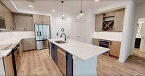 This well-appointed 740-square-foot, one-bedroom, one-bathroom apartment is designed with a modern aesthetic and thoughtful layout across a single floor. The open-concept kitchen and living area serves as the heart of the unit, featuring a large white quartz waterfall island with a black matte faucet and pendant globe lighting overhead. The kitchen is outfitted with flat-front light wood cabinetry, white quartz countertops, and a full suite of stainless steel appliances including a refrigerator, range, and microwave. A built-in wine rack and beverage cooler add a refined touch to the living area. Light wood-look luxury vinyl plank flooring runs seamlessly throughout the unit, complementing the bright white walls and clean trim. The 150-square-foot primary bedroom is bathed in natural light from large windows with roller shades, and includes a ceiling fan with integrated lighting. The spacious bathroom is a standout feature, showcasing a navy blue double vanity with LED-lit mirrors, a glass-enclosed walk-in shower with tile surround, a soaking tub, and large-format tile flooring. A dedicated closet completes this sleek, move-in-ready residence.