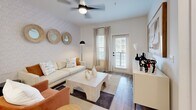 This single-story residence offers a thoughtfully designed open-concept layout that seamlessly connects the living, dining, and kitchen areas. The living room serves as a welcoming focal point, featuring wood-look plank flooring, a ceiling fan, and a textured accent wall adorned with a subtle scalloped pattern. Three decorative round mirrors and a statement arc floor lamp contribute to the space's contemporary aesthetic. A glass-panel door provides access to a private outdoor area, allowing natural light to filter through.

The kitchen is both functional and visually refined, equipped with gray shaker-style cabinetry, matte black hardware, white quartz countertops, and a classic white subway tile backsplash. Stainless steel appliances, including a refrigerator, range, and microwave, complement the space. A central island with seating for multiple guests and pendant globe lighting overhead makes it ideal for casual dining and entertaining.

The bedroom is a calm retreat with natural light from a window with blinds and neutral wall tones. Abstract artwork above the bed adds artistic character. The walk-in closet is outfitted with wire shelving systems and hanging rods, offering generous and organized storage space with carpeted flooring underfoot.