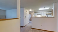 This single-floor residence offers a straightforward and functional layout designed for comfortable everyday living. The space features an open connection between the main living area and the kitchen, separated by a half-wall counter with warm wood-tone trim that provides a natural visual divider without closing off the floor plan. The kitchen is equipped with ample upper cabinetry in white with matching wood-tone accents, complemented by bright fluorescent overhead lighting that keeps the workspace well-illuminated. The flooring transitions from light-toned tile in the kitchen area to neutral carpet throughout the hallway and living spaces, adding warmth underfoot. The hallway extends toward the private rooms, creating a clear and organized flow through the home. Walls are finished in clean white throughout, lending a bright and airy feel to the interior. A textured ceiling adds subtle character to the space, while ceiling-mounted light fixtures provide consistent ambient lighting. An intercom panel is conveniently located along the hallway wall, adding a practical touch. Overall, the space presents a clean, move-in-ready environment with a simple, cohesive aesthetic that is both welcoming and easy to personalize.