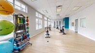 This dedicated exercise room offers a clean, open layout designed to accommodate a variety of fitness activities. The space is finished with light hardwood-style flooring that contributes to a bright, airy atmosphere, complemented by a drop ceiling with recessed fluorescent lighting panels that ensure consistent illumination throughout.

A row of large windows fitted with white horizontal blinds lines the perimeter walls, flooding the room with natural light and offering views of the surrounding outdoor greenery. The neutral gray walls are accented by a bold teal feature wall, which adds a vibrant, energizing touch to the overall aesthetic. A wall-mounted flat-screen television is positioned on the teal wall, making it ideal for guided workout sessions.

The room is equipped with multiple stationary spin bikes arranged in a row, providing ample space for cycling classes or individual use. Along one wall, a well-stocked equipment rack holds medicine balls, resistance bands, and other functional fitness accessories. The open floor plan allows for flexible use of the space beyond the installed equipment, supporting stretching, floor exercises, or additional fitness programming. A doorway connects to an adjacent room, enhancing the overall functionality of the facility.