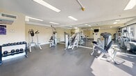 This fitness center is a comprehensively outfitted single-floor facility designed to support a full range of workout routines. The space is divided into distinct functional zones, each thoughtfully arranged to maximize usability and flow. The strength training area houses multiple cable and selectorized weight machines, including a lat pulldown, chest press, and seated row, complemented by a dedicated free weight station with a dumbbell rack. A large mirrored wall spans one side of the room, enhancing the sense of openness and allowing users to monitor their form during exercise. The cardio zone is well-stocked with treadmills, elliptical trainers, a stationary bike, and a Precor recumbent machine. A standout feature of this area is the expansive picture window that frames a direct view of a resort-style outdoor pool and surrounding palm-lined buildings, providing an energizing backdrop during workouts. Natural light floods this section of the room throughout the day. Overhead, recessed panel lighting illuminates the textured ceiling evenly across the entire space. Wall-mounted flat-screen televisions are positioned for visibility from multiple workout stations. The durable dark flooring throughout is practical and easy to maintain. The facility is accessible via glass-panel entry doors and includes posted fitness center rules for user guidance.