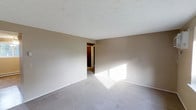 This single-floor space presents a spacious, open room finished with soft, neutral-toned walls that create a calm and understated atmosphere. The room is carpeted in a muted gray, adding warmth and comfort underfoot. A textured popcorn ceiling overhead provides a classic residential finish. Natural light streams in generously, casting bright patches across the floor and enhancing the airy feel of the room. The clean, unobstructed walls offer flexibility for a variety of furniture arrangements. An open doorway visible in the background leads to an adjoining area, suggesting additional rooms beyond and contributing to the overall sense of flow within the layout. White baseboards provide a crisp contrast against the neutral walls and carpet, lending a tidy, well-maintained appearance. The overall ambiance is bright, quiet, and move-in ready, making it a functional and adaptable space suited to a range of uses.