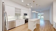 The Evergreen floor plan is a thoughtfully designed one-bedroom, one-bathroom residence that combines modern aesthetics with practical functionality. The open-concept kitchen and living area serves as the heart of the home, featuring light hardwood-style flooring, recessed lighting, and a ceiling fan that enhance the bright, airy atmosphere. The contemporary kitchen is outfitted with crisp white cabinetry, a tile backsplash, stainless steel appliances including a refrigerator with ice maker, range, microwave, and dishwasher, along with a central island topped with white countertops and illuminated by elegant brass pendant lights. Sliding glass doors at the end of the living area open directly to a private, fenced yard with lush green grass — a rare and desirable outdoor feature. The bedroom offers plush carpet flooring and connects to a spacious walk-in closet with built-in shelving and dual hanging rods, plus an additional closet for extra storage. The full bathroom is finished in a clean, all-white palette with a wide vanity featuring brass hardware, a lighted mirror, and tile flooring. A stacked washer and dryer is conveniently located within the unit, adding to the home's practical appeal.