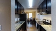 This two-bedroom, two-bathroom Magnolia residence is thoughtfully laid out across a single floor, offering a comfortable and functional living environment. The kitchen is a standout feature, designed in a galley style with rich, dark cabinetry that extends to the ceiling for generous storage capacity. Light granite countertops provide a striking contrast against the dark wood tones, while stainless steel appliances — including a full-size refrigerator and electric range — complete the space. A double-basin sink sits beneath under-cabinet lighting, adding both practicality and ambiance. The kitchen opens directly into a dedicated dining area, which is anchored by a classic chandelier and receives natural light through a well-positioned window, creating a bright and inviting atmosphere for meals. Warm hardwood-style flooring flows seamlessly from the kitchen through the dining space, lending continuity and a sense of warmth throughout. Textured ceilings and softly painted walls in a warm neutral tone contribute to the overall cozy yet polished character of the home. The layout is efficient and well-connected, making the most of the available space while maintaining a welcoming feel throughout.