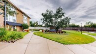 Located at 600 North Lilac Drive in Golden Valley, Minnesota, this outdoor amenity space offers a well-rounded set of recreational and functional facilities. The grounds are thoughtfully landscaped, featuring manicured lawns, decorative plantings, rock gardens, potted plants, and mature trees that provide natural shade along winding concrete walkways.

A central highlight is the outdoor swimming pool, enclosed by a white metal fence with a gated entry and surrounded by a spacious concrete deck furnished with lounge chairs — perfect for relaxing in warmer months. Adjacent to the pool is a covered patio area with a low ceiling, providing shade and shelter. This covered space includes seating and serves as a utility and storage area as well.

A shuffleboard court sits nearby, offering additional outdoor recreation options. The court is open-air and bordered by decorative planters, adding a touch of greenery to the space.

A cozy outdoor lounge area is set into the landscaping, featuring seating nestled among brick edging, stone accents, and lush shrubs — a pleasant spot for gathering beneath the shade of mature trees.

The space also includes a functional bathroom equipped with a pedestal sink, toilet, shower stall with curtain, and mosaic tile flooring. The multi-brick building facade visible from the grounds reflects a classic mid-century architectural style, grounding the amenity area within a well-established residential setting.