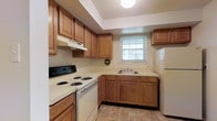 This single-floor space offers a practical and well-organized layout suited for comfortable everyday living. The kitchen is a standout feature, showcasing warm honey-toned wood cabinetry that lines the walls and provides generous storage. The cabinetry is complemented by light-colored countertops that extend across the full length of the workspace, offering ample room for meal preparation. A white electric range with coil burners anchors one side of the kitchen, paired with a matching white refrigerator on the opposite end, creating a cohesive look throughout. A double-basin sink is centrally positioned beneath a window fitted with horizontal blinds, allowing natural light to filter into the space while maintaining privacy. The kitchen floor features neutral stone-toned tiles that complement the overall color palette. An overhead range hood above the stove provides practical ventilation, while recessed step-down ceiling detailing adds a subtle architectural element above the main cooking area. A ceiling-mounted globe light fixture provides overhead illumination, contributing to the bright and functional atmosphere of the kitchen. The overall design is clean and utilitarian, prioritizing functionality and ease of use throughout the space.