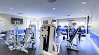This fitness center is thoughtfully designed to support a comprehensive workout experience. The single-level space is outfitted with an extensive selection of white-framed strength training machines, including cable systems, weight stations, and incline benches, arranged to allow comfortable movement throughout the floor. A dedicated cardio zone is visible toward the rear of the room, featuring treadmills and additional equipment positioned near large windows that invite ample natural light. The walls are finished in a clean white and accented with a bold blue stripe that adds a dynamic visual element to the space. Recessed ceiling lighting supplements the natural light, ensuring the room remains well-lit throughout the day. Mounted flat-screen televisions are positioned at multiple points along the walls, offering entertainment options during workouts. The flooring consists of durable dark rubber matting, providing safety and stability for a wide range of exercises. Posted signage, including fitness center policies, keeps the space organized and user-friendly. Overall, the gym presents a clean, modern, and energetic environment suitable for both strength and cardio training.