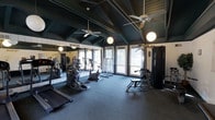 This fitness center is housed within a single-story structure distinguished by its vaulted, wood-beamed ceiling finished in deep green, which lends the space a distinctive and inviting character. Two ceiling fans circulate air throughout the room, complemented by globe pendant light fixtures that provide warm, even illumination. The flooring is covered in durable rubber matting, ideal for a high-use exercise environment. Large glass doors and windows along the far wall allow abundant natural light to fill the space while offering views of the surrounding outdoor landscape, creating an open and airy atmosphere. A wall of mirrors along one side visually expands the room and assists exercisers in monitoring their form. The space is equipped with a range of fitness machines, including treadmills, elliptical trainers, a stationary bike, and a cable strength training station, providing options for both cardiovascular and resistance training. The layout is thoughtfully organized to allow comfortable movement between equipment. The overall design balances a rustic architectural aesthetic with functional fitness amenities.