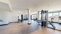 Located at 1450 Galapago St in Denver's vibrant Lincoln Park neighborhood, this fitness center offers a generously proportioned, single-floor layout designed to accommodate a full range of workout activities. The space is characterized by soaring ceilings and an abundance of natural light, courtesy of expansive floor-to-ceiling windows that frame views of the surrounding Denver cityscape.

The open floor plan is thoughtfully organized into distinct training zones. A dedicated cardio area lines the windowed perimeter, housing a full row of treadmills, elliptical machines, and stationary bikes — all positioned to take advantage of the outdoor views. The strength training section features multiple cable and multi-functional machines, weight benches, and a well-stocked free weights area complete with kettlebell racks and a comprehensive dumbbell selection displayed along mirrored walls.

Structural columns punctuate the space while maintaining an airy, unobstructed feel throughout. The flooring transitions between durable rubber and low-pile carpet, providing appropriate surfaces for various exercise types. Accent walls — including a bold teal feature wall and a decorative yellow patterned panel — add visual interest against the otherwise clean, white interior. Recessed and pendant lighting complement the natural light, ensuring the space remains bright and energizing throughout the day.