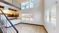 Located in Flagstaff, Arizona, this two-story, two-bedroom, two-bathroom residence combines modern design with functional living across two well-proportioned floors. The main level opens to a dramatic living area distinguished by soaring double-height ceilings and an abundance of natural light pouring through large windows, which frame views of the surrounding mountains and landscape. Light wood-look flooring and dark baseboards run consistently throughout, lending a clean, contemporary aesthetic. The kitchen is thoughtfully appointed with rich dark wood cabinetry, granite-style countertops, stainless steel appliances including a refrigerator, range, and microwave, and pendant lighting beneath an exposed wood beam accent — a striking architectural detail that adds warmth and character. A sleek metal cable railing staircase leads to the upper level, where both bedrooms are situated. The spacious primary bedroom features vaulted ceilings, multiple windows with mountain views, and a sliding closet door. Both bathrooms are finished with wood-toned vanities, granite-style countertops, and wood-look tile flooring. One bathroom offers a walk-in shower, while the other features a full shower-tub combination. The upper hallway overlooks the main living area, reinforcing the open, airy feel of this well-designed Flagstaff home.